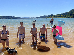 A group of people standing on the shore of a lake in bathing suits on a sunny day.